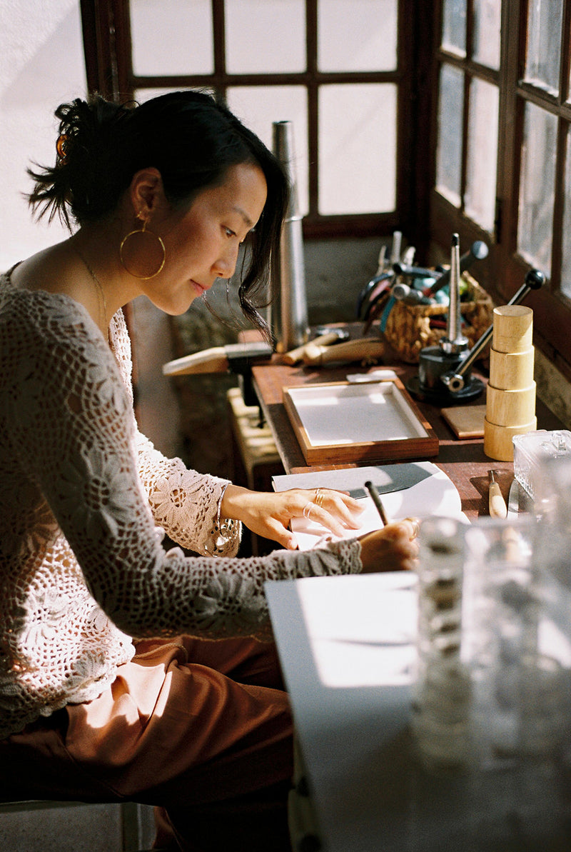 Woman sitting at a jewelers bench drawing a design in a notebook.